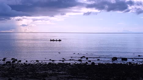 Un-Grupo-De-Pescadores-Locales-Con-Siluetas-Remando-En-Un-Barco-De-Pesca-Tradicional-De-Madera-Sobre-El-Arrecife-De-Coral-Con-Un-Hermoso-Amanecer-De-Neblina-Púrpura-En-La-Isla-De-Atauro,-Timor-Leste
