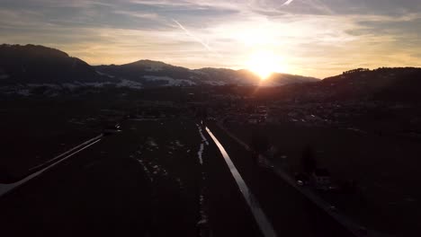 Beautiful-aerial-flight-over-a-road-and-a-village-while-sunset-in-Switzerland