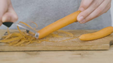 Zwei-Hände-Schälen-Eine-Karotte-Mit-Einem-Kartoffelschäler-Auf-Einem-Holzschneidebrett,-Eine-Karotte-Wurde-Bereits-Geschält