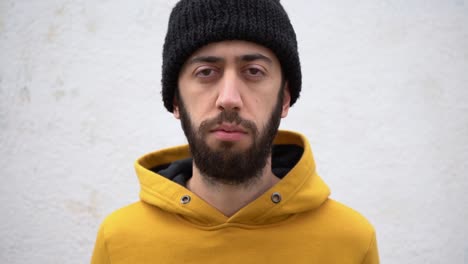 Bearded-Man-With-Serious-Face-In-Mustard-Hoodie-And-Knitted-Beanie-Pointing-Finger-At-His-Eye-Showing-To-Someone-I-Am-Watching-You-Standing-Against-A-Concrete-Wall