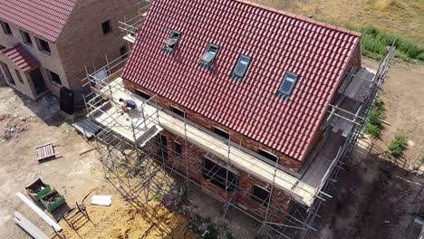 Rotating-aerial-view-of-a-scaffolded-new-build-house-under-construction,-showing-the-front-elevation-and-the-gable-end