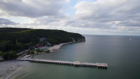 Orlowo-pier-and-public-beach-coastline-on-summer-cloudy-day-in-Gdynia,-Poland---Aerial-uprise-reversing-shot