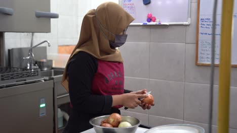 Muslim-lady-house-wife-wearing-hijab-headscarf-working-in-the-kitchen,-peeling-brown-onions-with-paring-knife