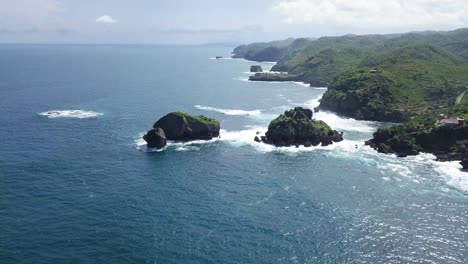 Aerial-view-of-coral-island-with-overgrown-by-trees-in-ocean,hit-by-waves-during-sunny-day---TIMANG-ISLAND,-YOGYAKARTA,-INDONESIA