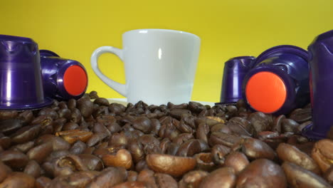 White-mug-with-roasted-coffee-beans-and-plastic-coffee-capsule-in-front-of-yellow-screen