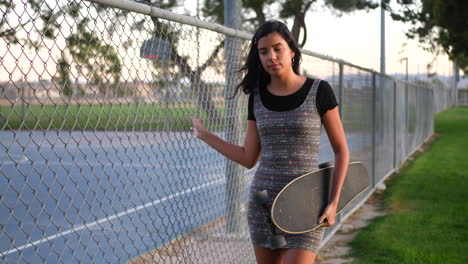 A-beautiful-young-hispanic-woman-walking-through-an-urban-city-park-with-her-skateboard-along-a-chainlink-fence-SLOW-MOTION