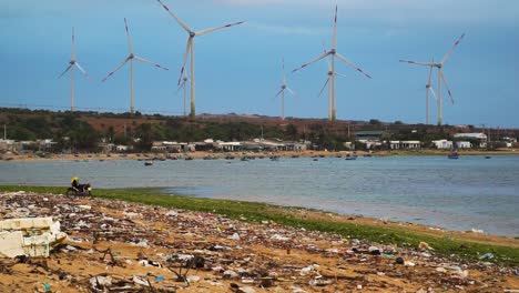 Rotierende-Rotorblätter-Eines-Sauberen-Und-Erneuerbaren-Windkraftparks-Im-Hintergrund-Und-Verschmutztes-Strandufer-Mit-Plastik,-Müll-Und-Schmutz