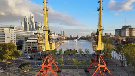 Cinematic-aerial-flying-between-two-old-port-cranes-and-revealing-Puerto-Madero-waterfront-skyline