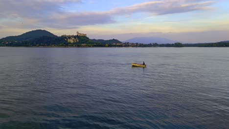 Fisherman-rowing-wooden-fishing-boat-on-Maggiore-lake-in-Italy-with-Angera-fortress-in-background