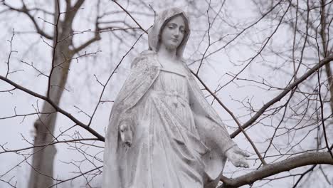 Weiße-Marmorsteinstatue-Der-Mutter-Maria-An-Einem-Düsteren-Tag-Mit-Wehenden-Ästen-Im-Hintergrund