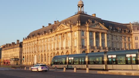 Place-De-La-Bourse-In-Bordeaux-Bei-Sonnenaufgang-Mit-Vorbeifahrender-Straßenbahn-Und-Einigen-Autos,-Mit-Dem-Französischen-Zollmuseum-Im-Hintergrund