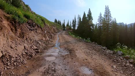 POV-following-a-Jeep-driving-up-Alpine-Loop-trail-cut-into-hillside,-past-pine-trees-in-San-Jan-Mountains-near-Silverton-Colorado