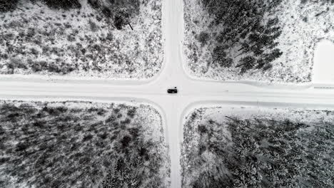 Aerial-bird's-eye-view-of-a-black-car-standing-in-the-middle-of-the-crossroad-with-snow-covered-on-roads-and-pine-tree-forest-on-all-four-sides