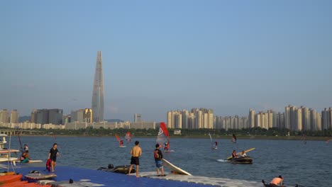 Korean-People-walking-on-the-floating-pier-when-others-Windsurfing-on-Han-river-next-to-Lotte-World-Tower,-Seoul-on-Sunset