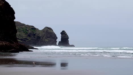 Piha-Beach-black-beach,-rocky-outcrop-and-ocean-waves-on-a-grey,-overcast-rainy-winters-day-in-Auckland,-New-Zealand