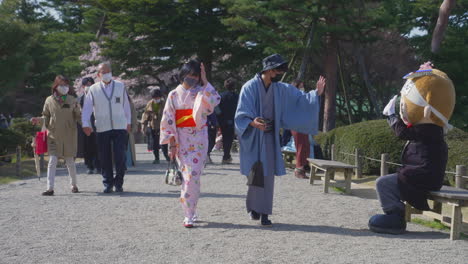 Junges-Asiatisches-Japanisches-Maskiertes-Paar-Im-Kimono,-Das-Im-Kenrokuen-Garten-Spaziert-Und-Einem-Lokalen-Maskottchen-Litakun-Zuwinkt