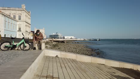 Älterer-Mann-Sitzt-Auf-Einer-Mauer-Am-Ufer-In-Lissabon-Mit-Klarem-Blauen-Himmel