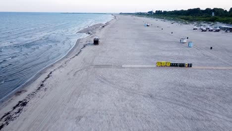 Aerial-view-of-garbage-truck-moving-along-the-empty-beach-in-early-summer-morning-after-the-sunrise,-preparing-to-load-waste,-wide-angle-drone-shot-moving-forward