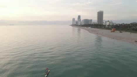 Luftaufnahmen-überholen-Einsamen-Paddleboarder-Am-Sandstrand,-Als-Miami-Aufwacht