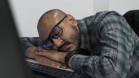 A-young-Indian-entrepreneur-slumped-over-an-office-desk,-asleep-in-front-of-his-computer-exhausted-from-working-late-putting-together-his-business-proposal