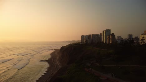 Aerial-view-of-seaside-buildings-on-the-coastline-of-Lima-city,-hazy-sunset-sky