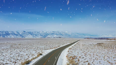 Majestic-iconic-country-road-with-mountain-range-in-horizon-during-snowfall,-aerial-drone-view