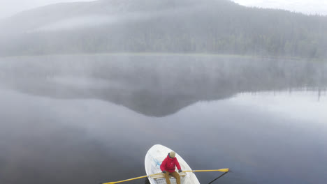 Aerial-view-of-going-over-a-person-rowing-a-boat-on-a-foggy-lake-with-forest-and-mountain-in-the-landscape