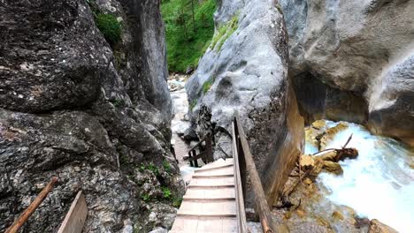 Smooth-POV-footage-of-someone-walking-down-wooden-stairs-between-two-rocks,-next-to-a-wild-white-river-in-a-beautiful-canyon