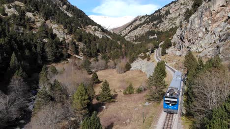 Aerial:-blue-rack-train-in-the-Pyrenees-mountains-going-close-to-the-river-and-across-a-very-narrow-valley