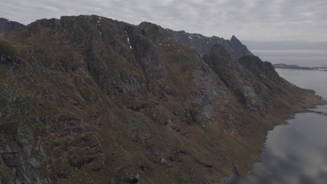 Aerial-drone-view-flying-parallel-to-a-steep,-rocky-mountain-wall-in-cloudy-Lofoten,-Norway