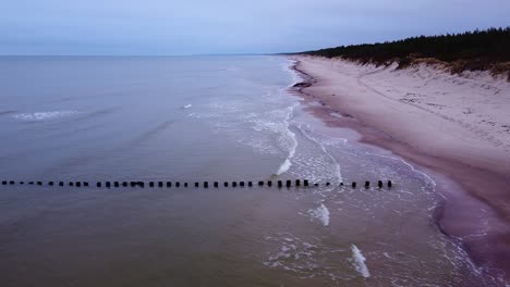 Beautiful-aerial-view-of-an-old-wooden-pier-at-the-Baltic-sea-coastline,-overcast-day,-white-sand-beach-affected-by-sea-coastal-erosion,-calm-seashore,-ascending-wide-angle-drone-shot-moving-left