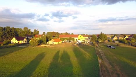 Ruhigerer-Luftdrohnenflug,-Vorwärts-Fliegender-Drohnenschuss-Von-Einem-Bauernhof-Mit-Langen-Schatten-Von-Bäumen-Bei-Sommerlichem-Sonnenuntergang-An-Einem-See-In-Einem-Kleinen-Dorf-In-Brandenburg,-Deutschland