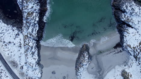 aerial-birdseye-shot-of-Andoya-beach-Norway,-with-beautiful-contrast-between-the-snow-covered-rocks,-green-sea-and-wave