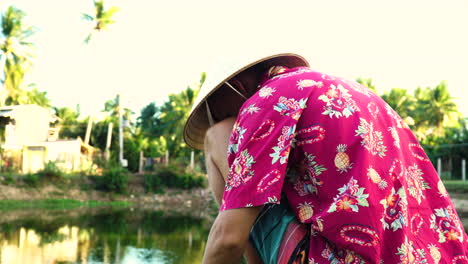 Man-with-exotic-shirt-and-conical-hat-preparing-to-fish-in-local-water-pond,-sunny-tropical-evening