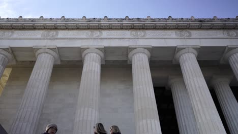 Tourists-on-Lincoln-Memorial,-Low-angle-panning-through-Columns-against-blue-sky
