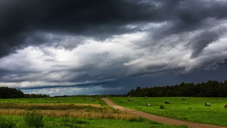 Shot-of-mystic-dark-clouds-flying-over-green-field-with-hay-bales-beside-a-gravel-pthway-in-timelapse