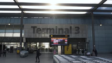 Reihe-Von-Gepäckwagen-Vor-Dem-Heathrow-Terminal-3-Gebäude-Mit-Abflugtafel-Der-Fluggesellschaft-Und-Sonne,-Die-Durch-Den-Dachüberstand-Scheint