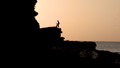 Man-with-fishing-pole-over-large-ocean-rocky-cliffs-at-the-beach-during-sunset,-Handheld-medium-shot