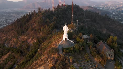 Aerial-dolly-in-over-statue-in-Sanctuary-of-the-Immaculate-Conception-in-San-Cristobal-Hill-top,-Santiago-city-in-background-at-sunset,-Chile
