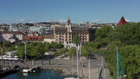Drone-flying-over-yacht-harbor-towards-clock-tower-in-downtown-Lausanna,-Switzerland