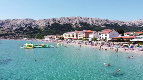 Baska-Beach,-Krk-island,-Croatia---Aerial-Drone-View-of-the-Boulevard,-Tourists,-Sunbeds-and-Water-Playground-during-Summer-Holiday
