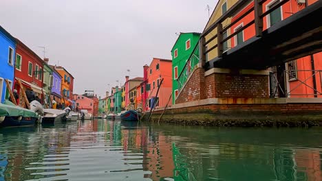 Water-surface-pov-view-of-Burano-picturesque-colored-houses-and-canal-with-moored-boats,-Italy