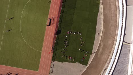 Eine-Dynamisch-Rotierende-Luftaufnahme-Einer-Gruppe-Von-Schülern,-Die-Auf-Dem-Rasen-Des-Leichtathletikstadions-Liegen-Und-Übungen-Und-Armbewegungen-Machen
