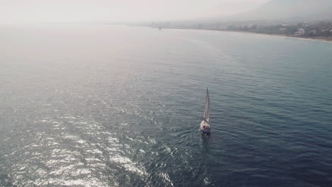 Yacht-sailing-along-the-coastline-of-Spain,-drone-moving-backwards-and-tilting-into-the-horizon