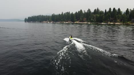 Drone-shot-following-a-person-driving-a-jet-ski-at-Payette-Lake-in-McCall,-Idaho-with-the-shore-and-trees-in-the-background