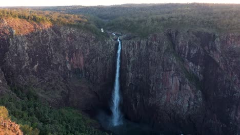 Wallaman-Falls-in-Queensland-dusk-aerial-view-moving-backwards,-tall-waterfall-with-golden-sunlight