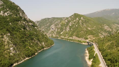 Aufsteigende-Luftaufnahme-über-Einen-Breiten-Teil-Des-Flusses-Neretva-Mit-Einer-Brücke-über-Einen-Nebenfluss-In-Bosnien-Und-Der-Straße-Am-Fuße-Der-Berge