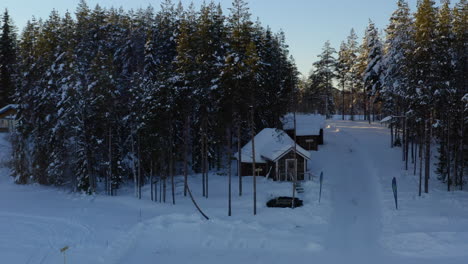 Luftaufnahme,-Vorbei-An-Einer-Abgelegenen,-Schneebedeckten-Waldhütte-Im-Winter,-Die-Sich-Bei-Sonnenaufgang-über-Alpenbäumen-Erhebt