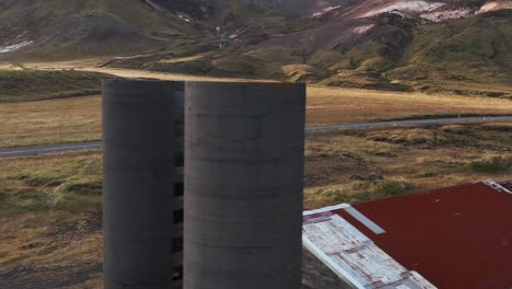 Abandoned-broken-building-with-grain-silos-in-Icelandic-valley,-white-car-passing-by-in-background,-aerial