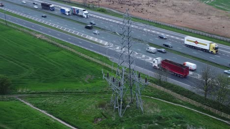 Vehicles-on-M62-motorway-passing-pylon-tower-on-countryside-farmland-fields-aerial-view-descent-from-high-angle